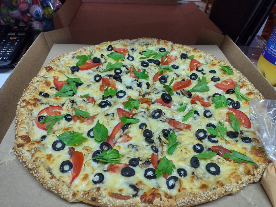 Pizza de tamaño grande con aceitunas negras, queso, tomate y perejil en caja de cartón.