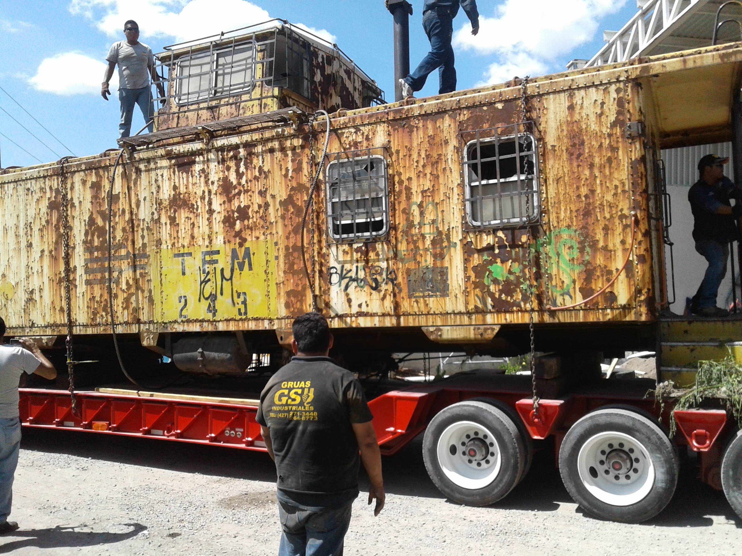 Antiguo vagón de tren oxidado siendo transportado en un remolque rojo, con personas trabajando alrededor.