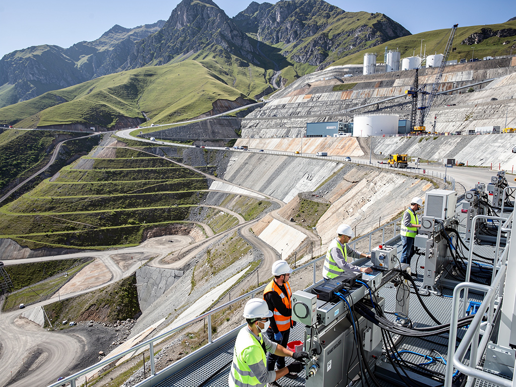 Ingenieros con cascos trabajando en equipos industriales en una obra de presa hidroeléctrica en montañas verdes