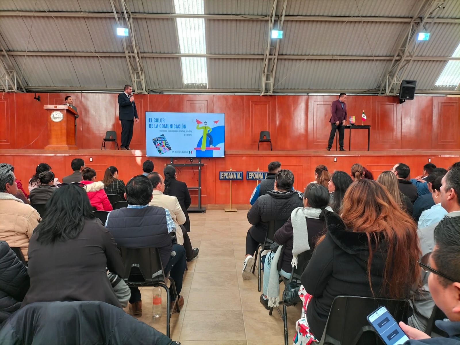 Estudiantes en un gimnasio universitario durante una feria académica con mesas de registro, pantalla de presentación y gran asistencia Estudiantes en un gimnasio universitario durante una feria académica con mesas de registro, pantalla de presentación y gran asistencia