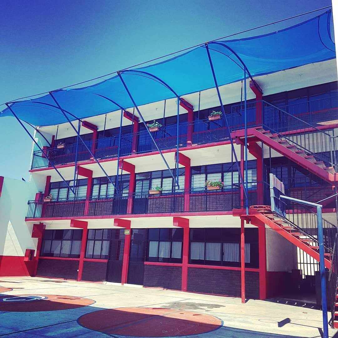 Edificio escolar de tres pisos con escaleras rojas y toldo azul en día soleado.