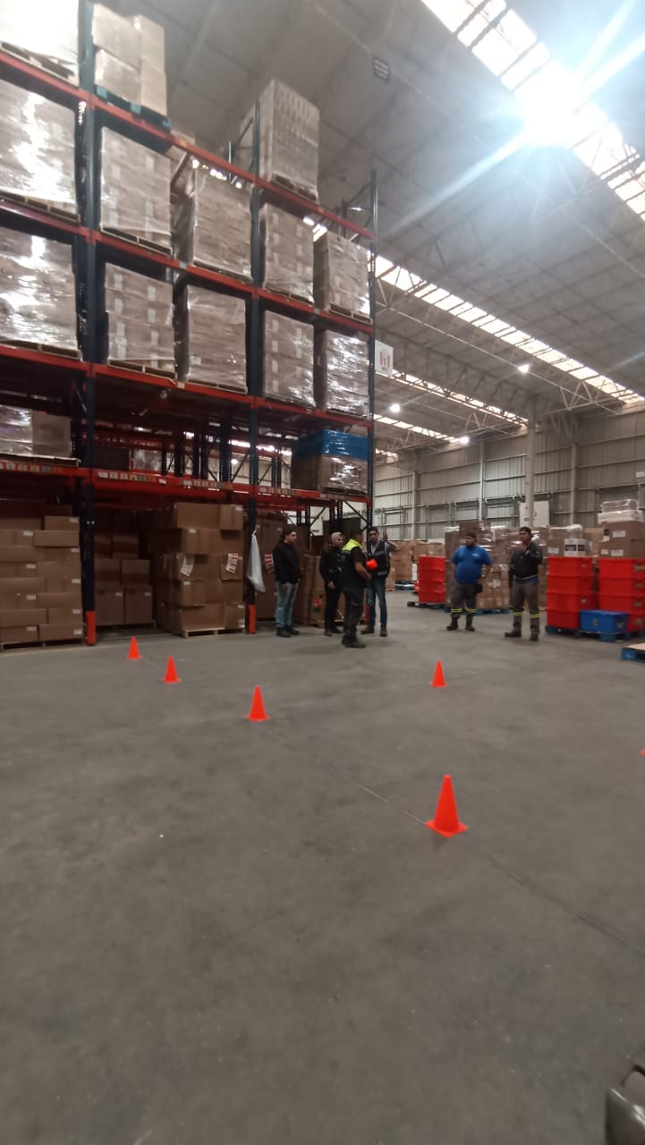 Interior de almacén con estanterías altas, cajas apiladas y un grupo de personas discutiendo frente a conos naranjas.