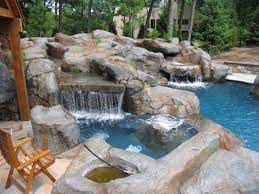 Luxurious outdoor swimming pool area with natural rock waterfalls, surrounded by lush greenery and wooden deck seating.