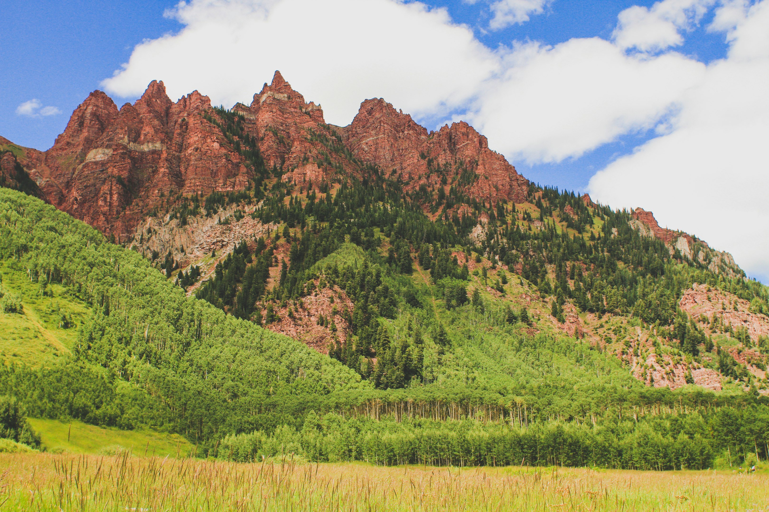 Scenic view of green forested mountain with red rock formations under blue sky with clouds.