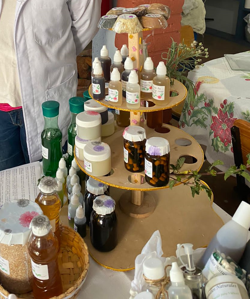 Mesa de exposición con productos naturales en frascos, incluyendo aceites y cremas, sobre un soporte de madera junto a una persona con bata blanca.