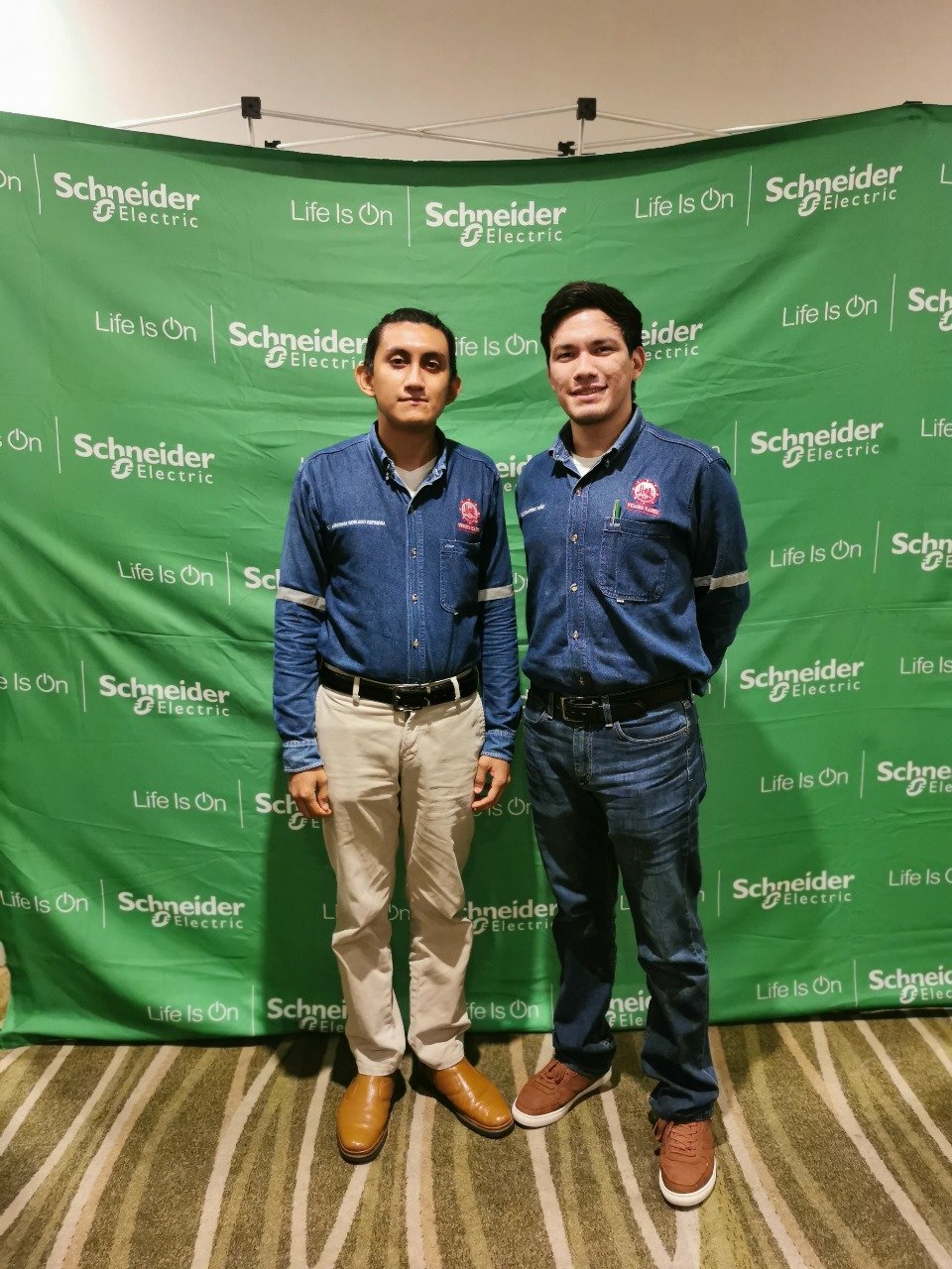 Dos hombres con camisas de mezclilla frente a un fondo verde de Schneider Electric con las palabras 'Life Is On'. Dos hombres con camisas de mezclilla frente a un fondo verde de Schneider Electric con las palabras 'Life Is On'.