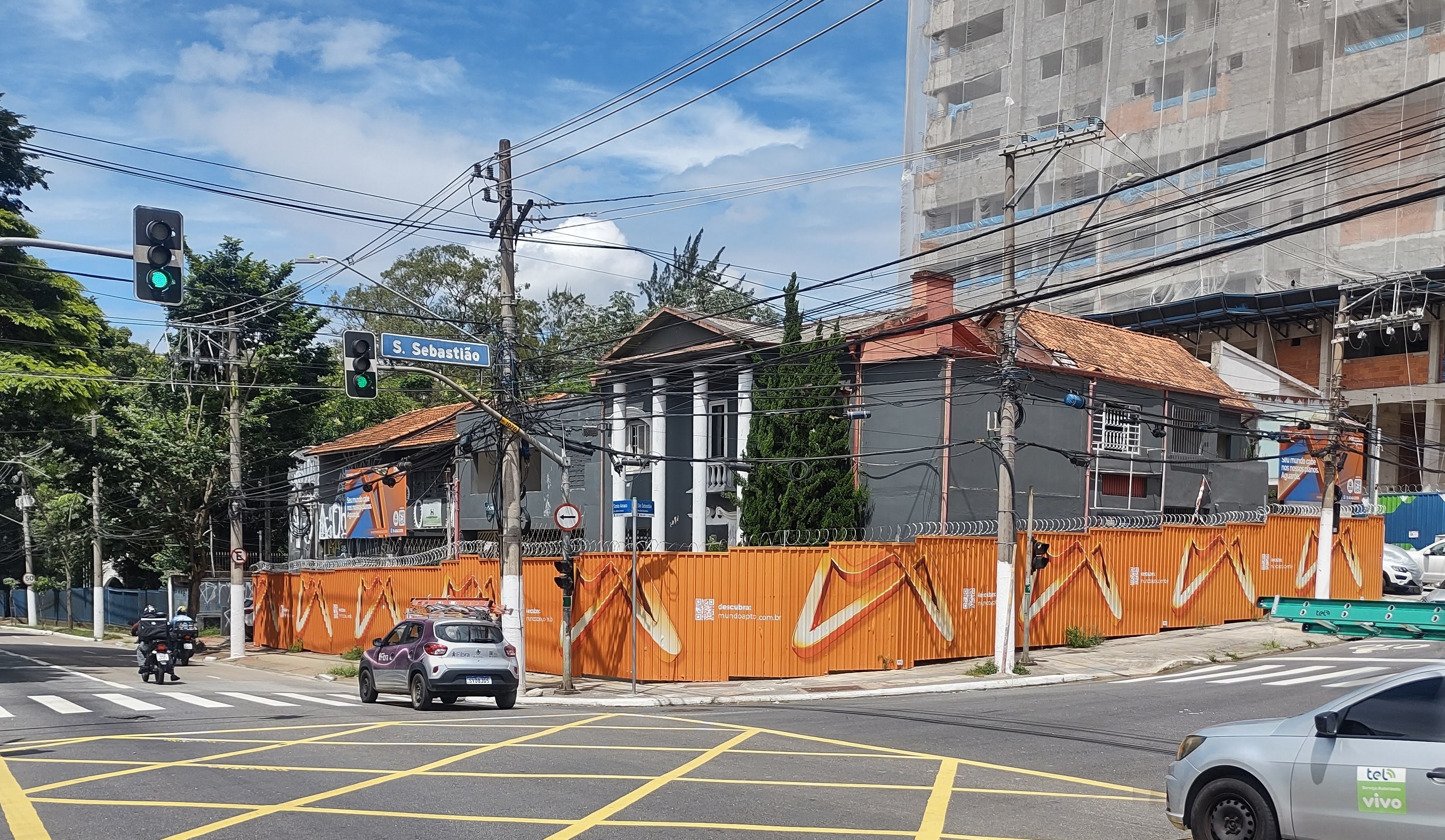 Cruzamento em São Sebastião com construção cercada por tapumes laranja, sinal de trânsito verde, e edifício ao fundo.
