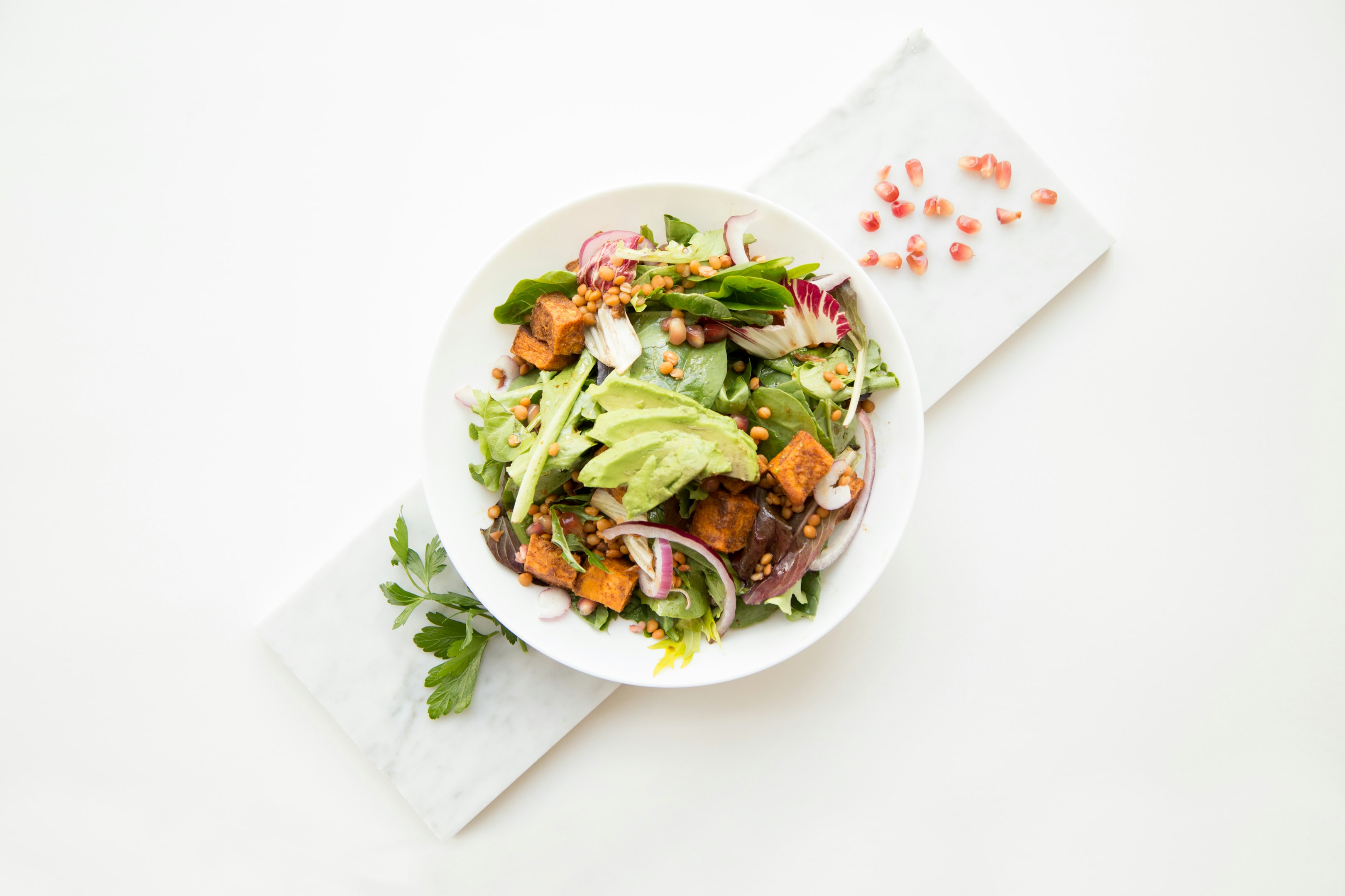 Fresh salad with avocado, leafy greens, roasted sweet potatoes, red onion, and pomegranate seeds on a white marble background.