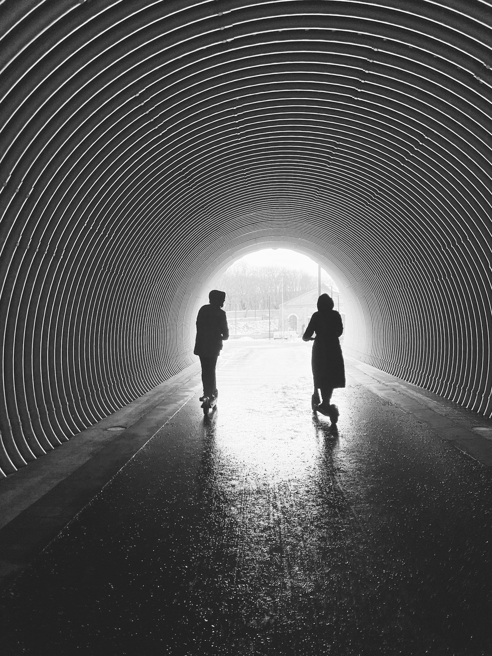 Siluetas de dos personas caminando en un túnel iluminado al fondo con un patrón de líneas curvas.
