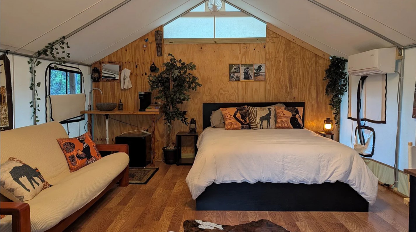 Luxury glamping tent on a wooden deck at sunset, surrounded by scenic landscape with trees and softly lit sky.