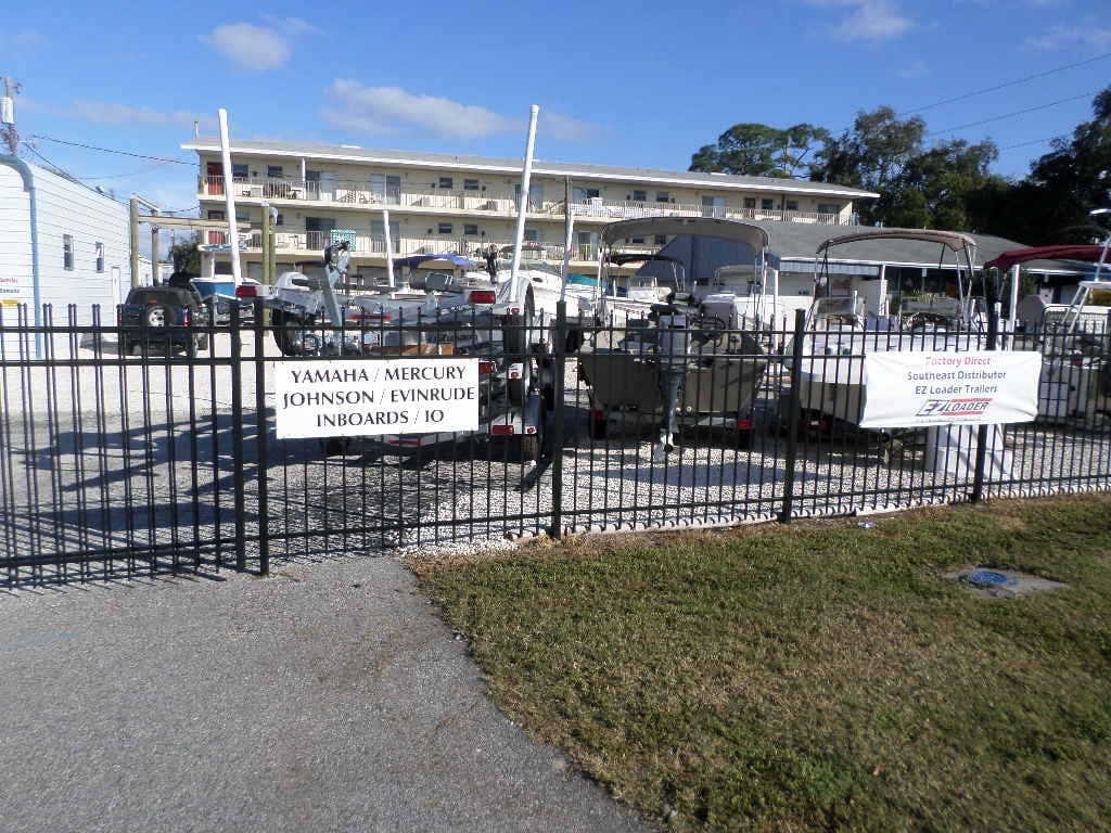 Boat and trailer sales lot with outboard motor service signs behind black metal fence