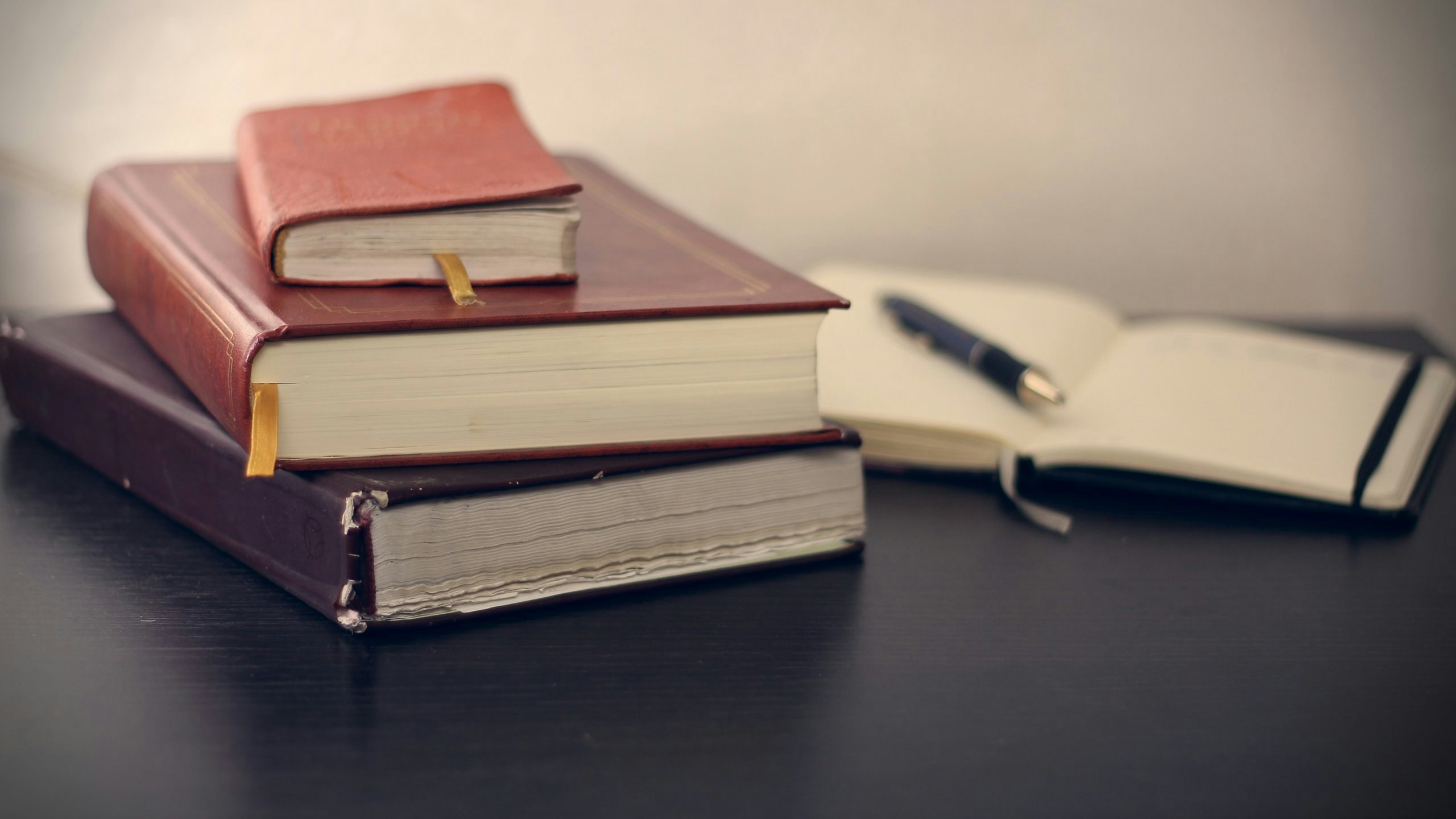 Stack of hardcover books with bookmarks on a dark table beside an open notebook and pen.