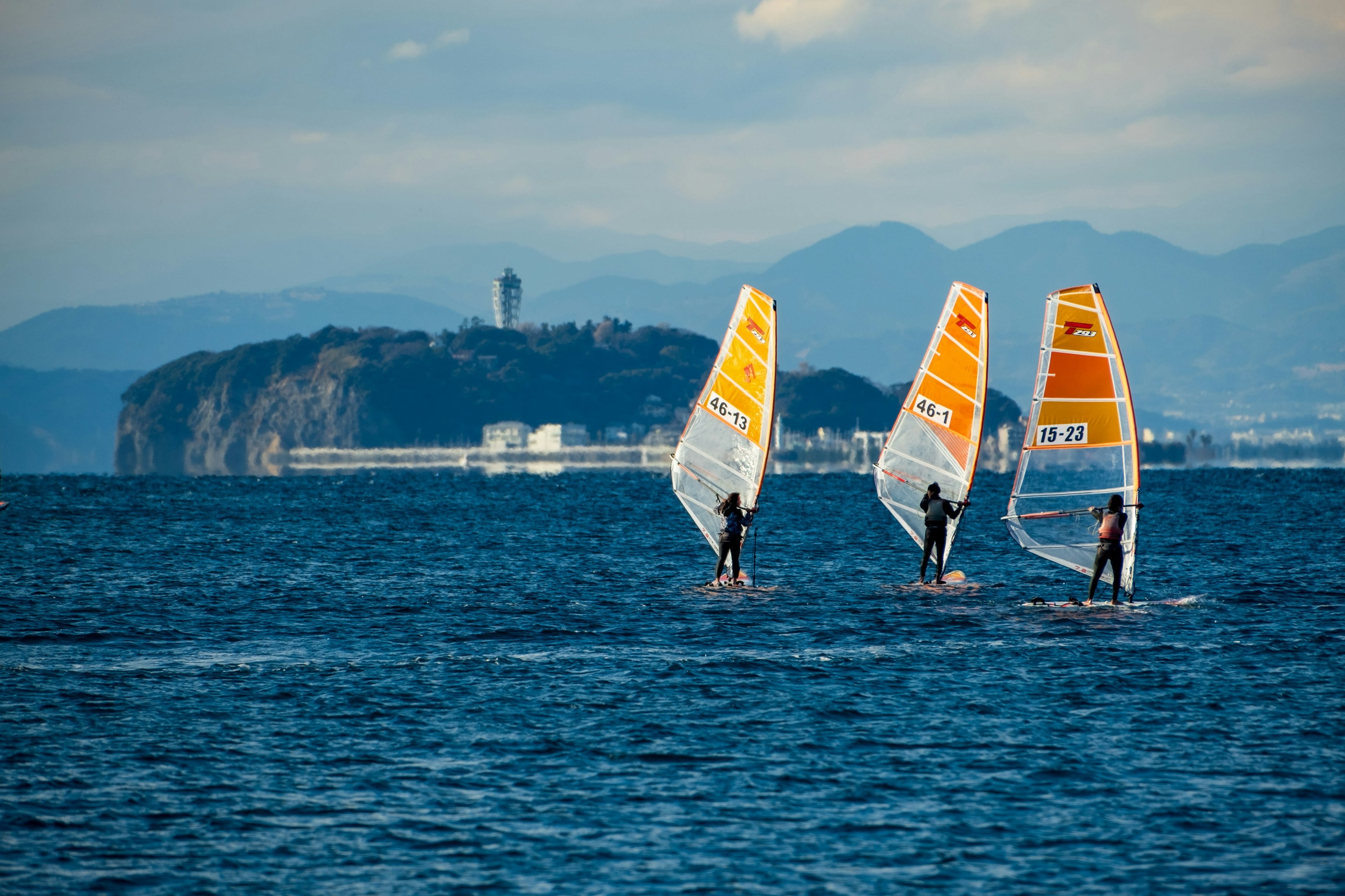 Three windsurfers with orange sails glide across a blue sea with a coastal landscape and mountains in the background. Three windsurfers with orange sails glide across a blue sea with a coastal landscape and mountains in the background.