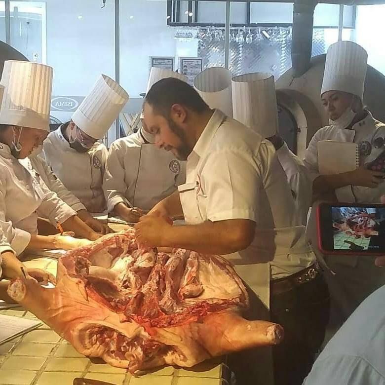 Grupo de estudiantes de cocina observa a un chef mientras realiza un corte de carne de cerdo en una clase práctica.