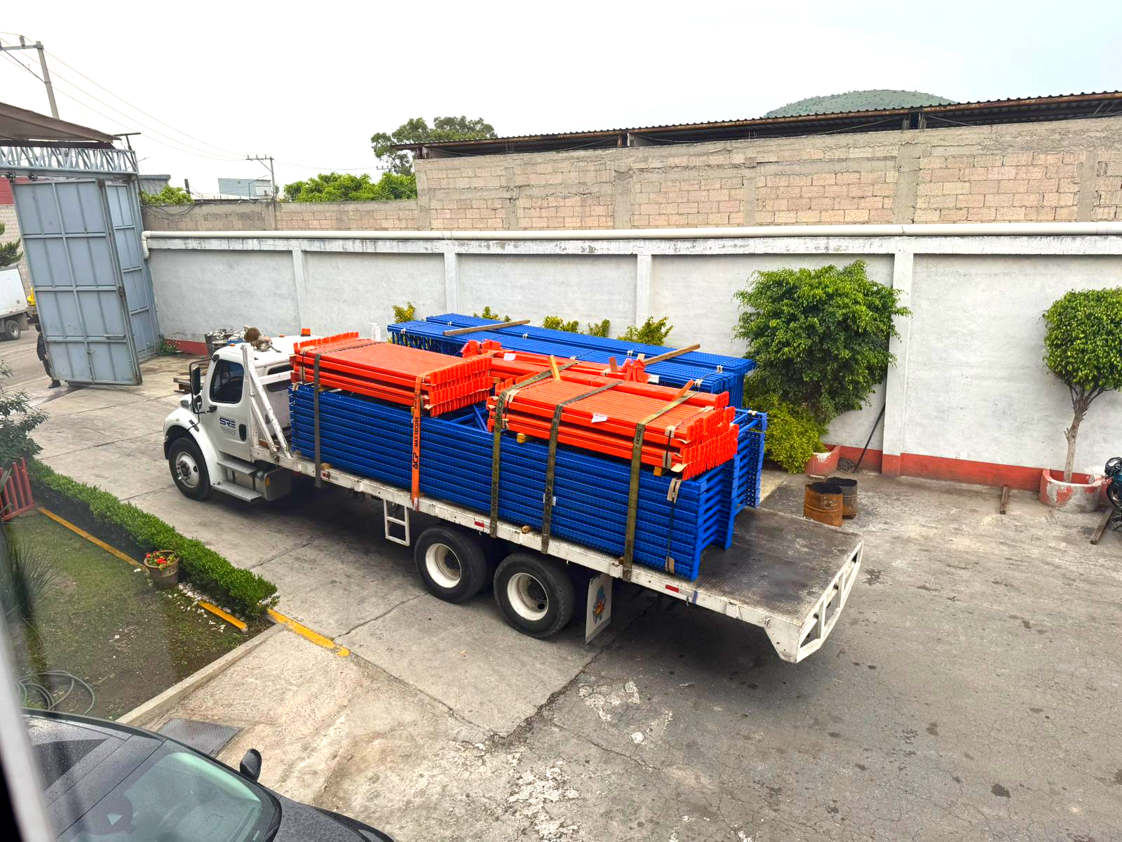 Camión de plataforma transportando estructuras metálicas azules y naranjas en un patio industrial.