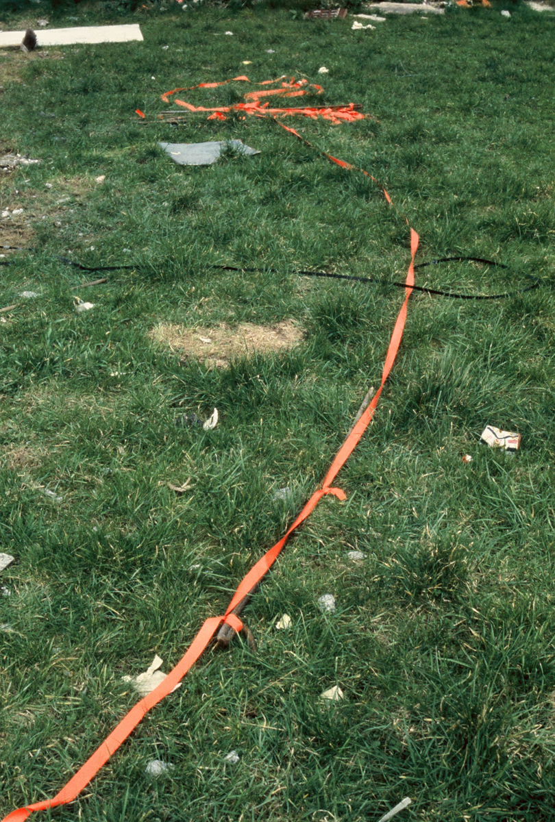 A long piece of orange barrier tape and other trash on grassy green wasteland.