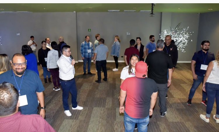 Grupo de personas sonriendo mientras sostienen certificados de reconocimiento, en un ambiente industrial con letreros de bienvenida en inglés y español.