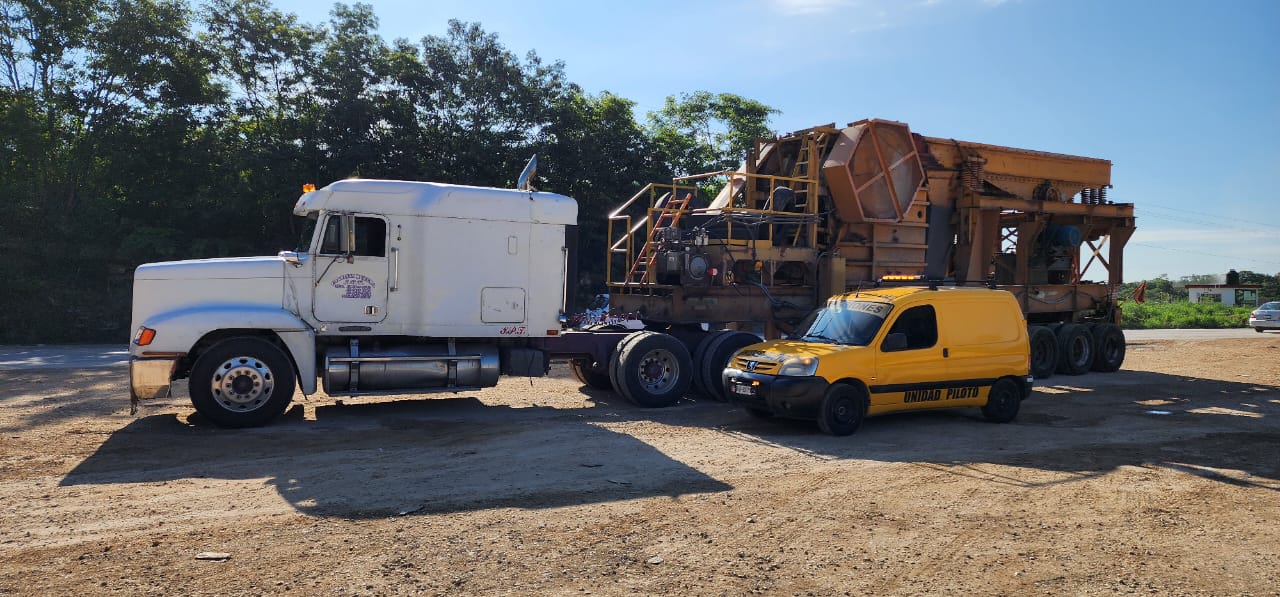 Camión blanco y vehículo de asistencia amarillo estacionados en terreno arenoso junto a maquinaria pesada, con árboles al fondo bajo un cielo despejado.