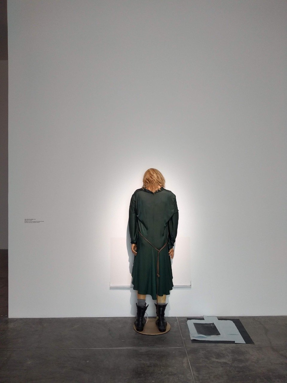 Maniquí con peluca rubia y vestido verde oscuro frente a una pared blanca en una galería de arte.