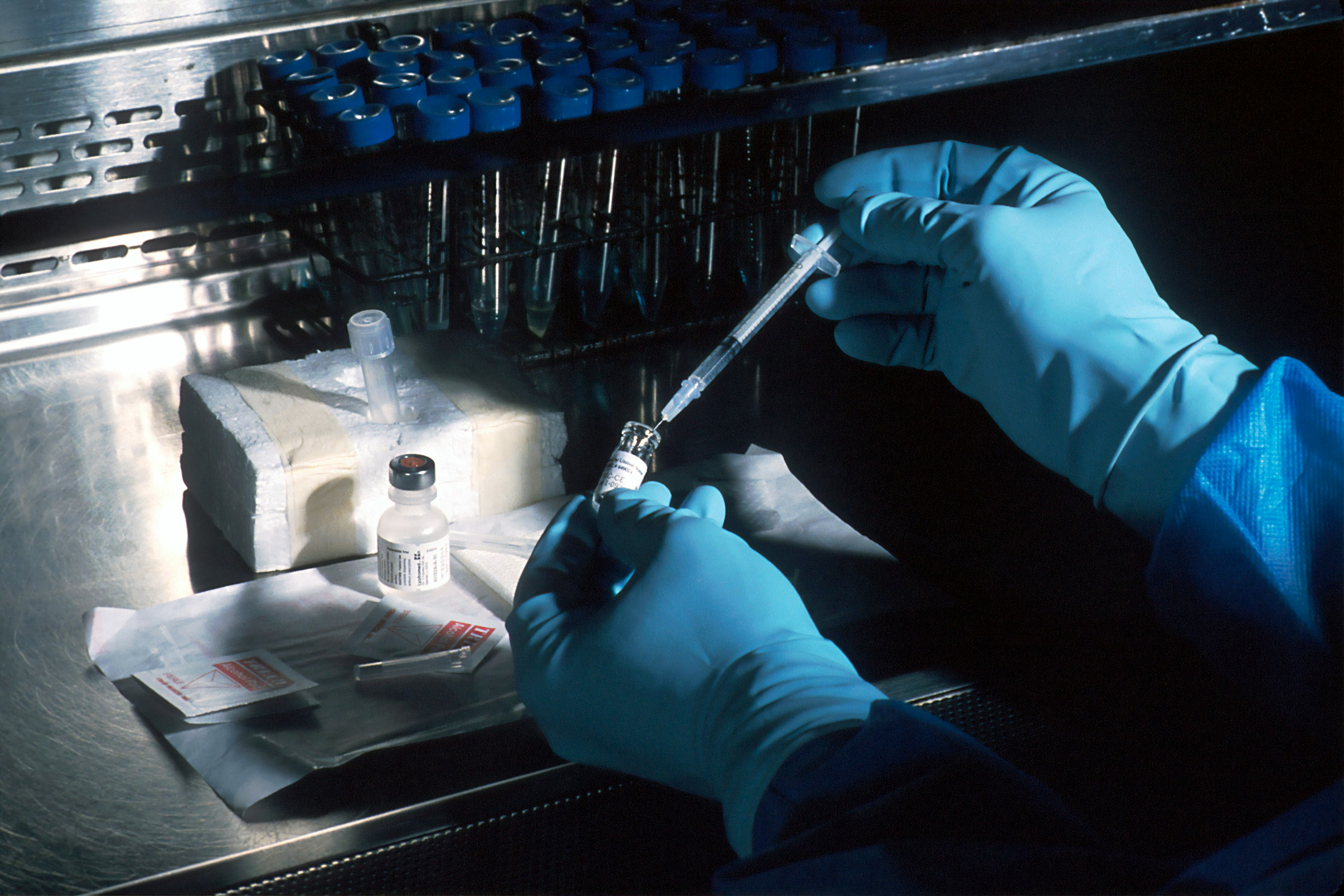 Investigadora en laboratorio con bata blanca, gafas protectoras y guantes azules, sosteniendo un tubo de ensayo.
