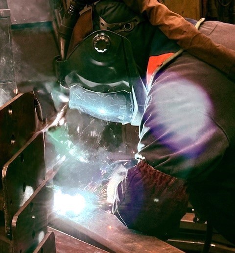 Welder in protective gear working on metal in an industrial workshop, surrounded by tools and machinery.