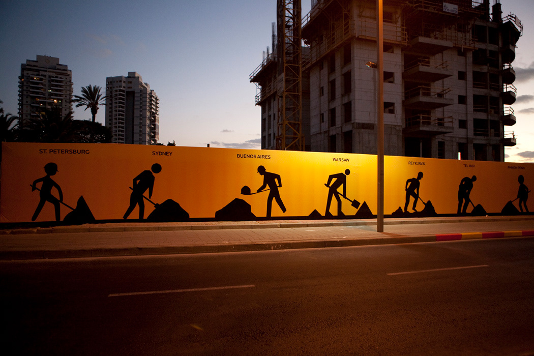 Mural iluminado à noite com silhuetas de trabalhadores e nomes de cidades, construção ao fundo.