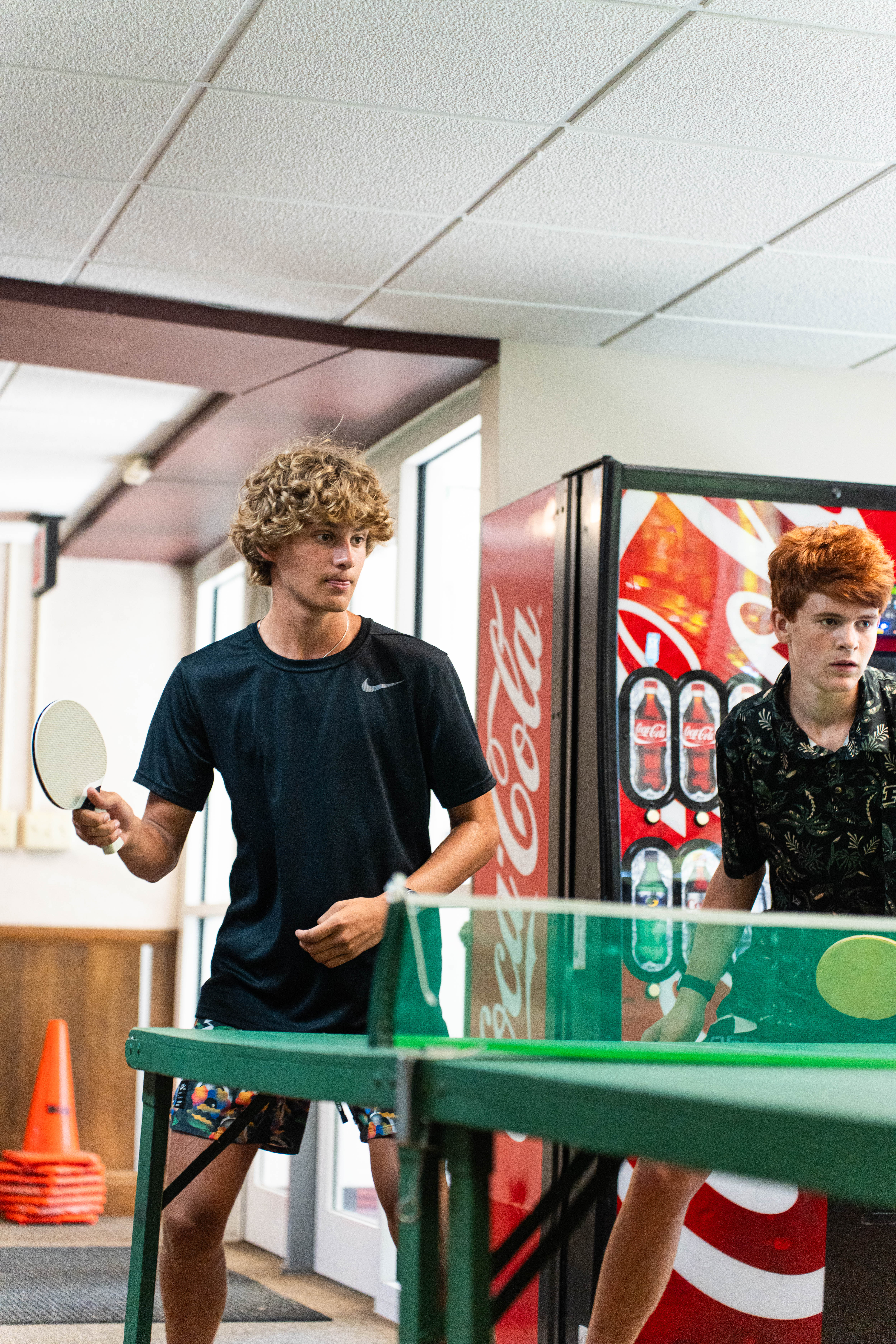 Two young men playing table tennis indoors near a Coca-Cola vending machine.