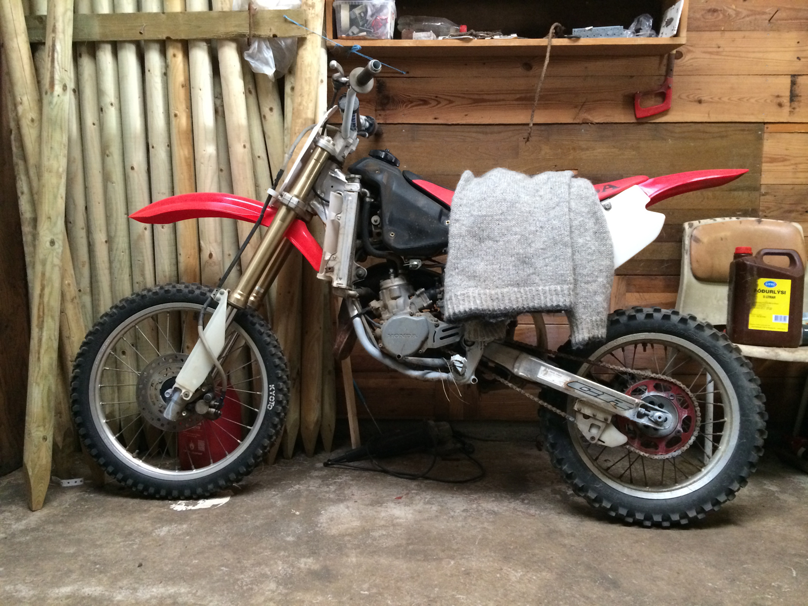A motocross motorcycle, a sweater on the seat, leaning on a wall with staves.