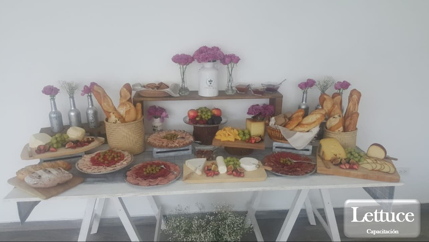Mesa de charcutería con variedad de quesos, embutidos, frutas y panes, decorada con flores y botellas de vidrio.