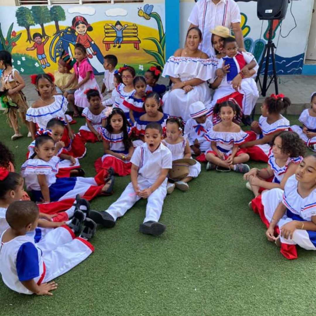 Niños en un evento cultural, vestidos con trajes tradicionales coloridos, sentados sobre césped artificial frente a un mural colorido, sonriendo y disfrutando el momento. Niños en un evento cultural, vestidos con trajes tradicionales coloridos, sentados sobre césped artificial frente a un mural colorido, sonriendo y disfrutando el momento.