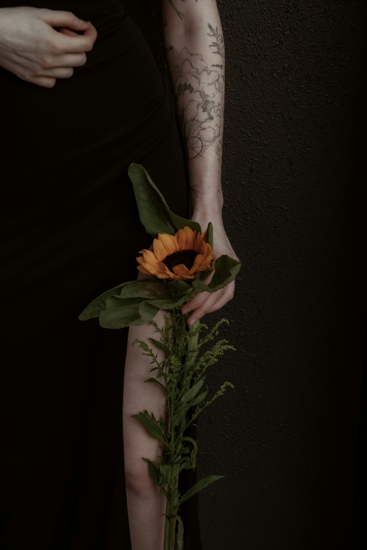 Woman with floral tattoos, long dark hair, in a black top, standing in front of shelves with photos and plants.
