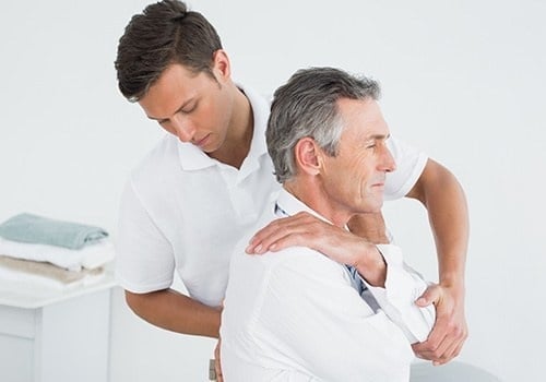 Young male physiotherapist assisting senior male patient with shoulder exercise in clinic setting.