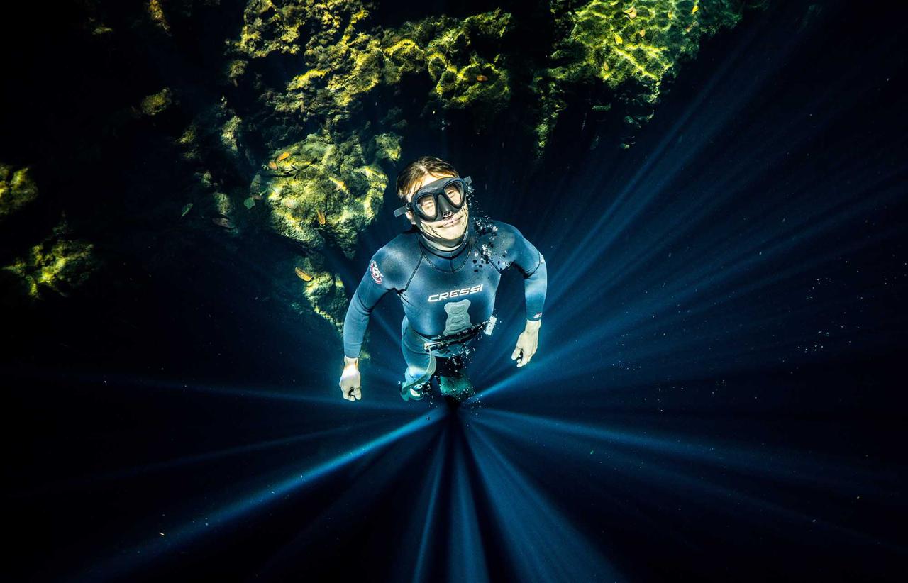 Piscina al aire libre rodeada de palmeras y edificios lujosos con sillas de playa, bajo un cielo azul con nubes y montañas al fondo.