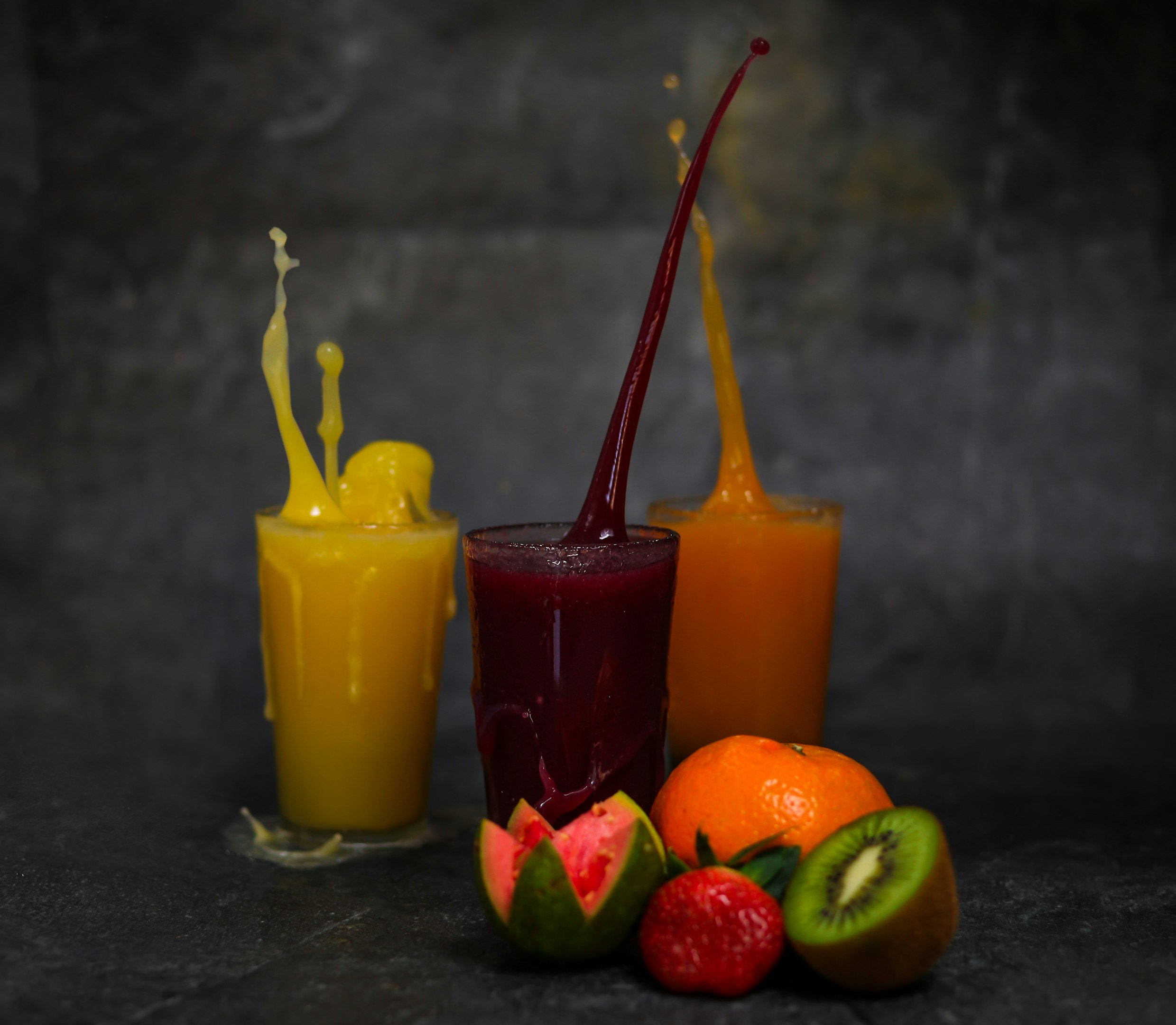 Tres vasos de jugos de frutas coloridos con salpicaduras y frutas frescas delante, incluyendo kiwi, guayaba, fresa y naranja.