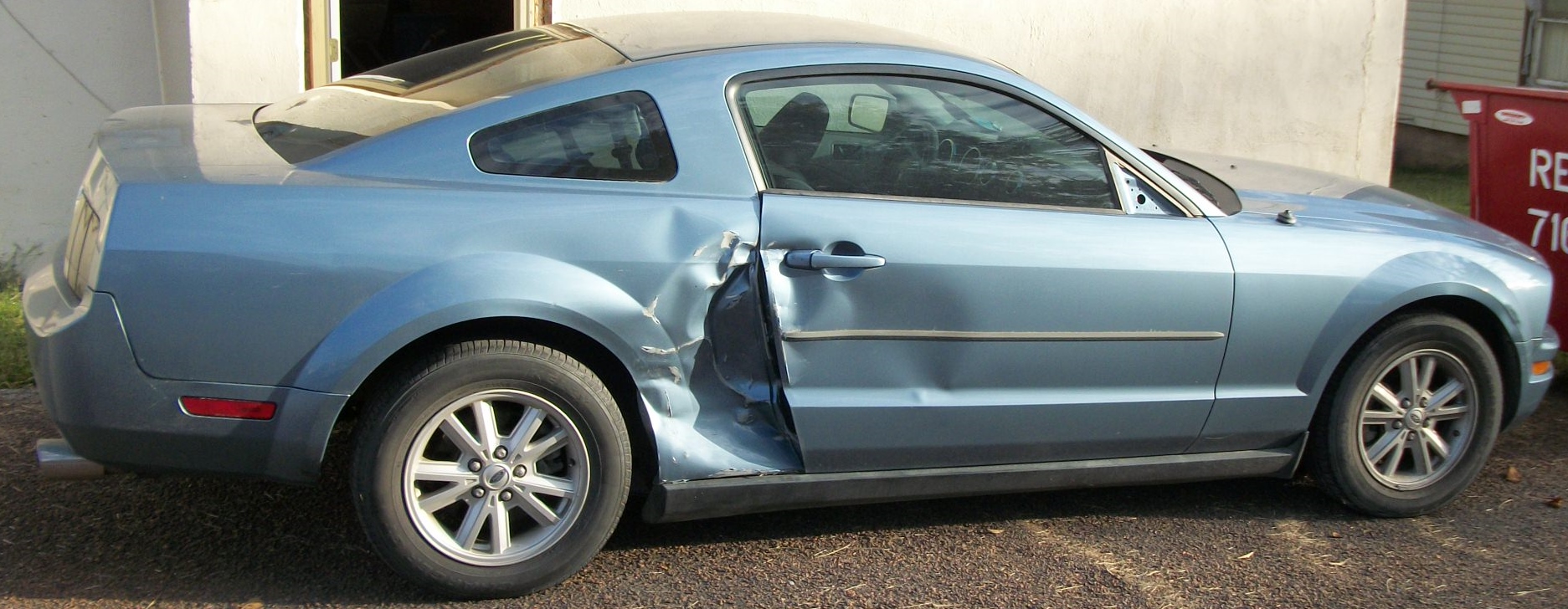 Damaged blue car with dented side parked on asphalt near white building.