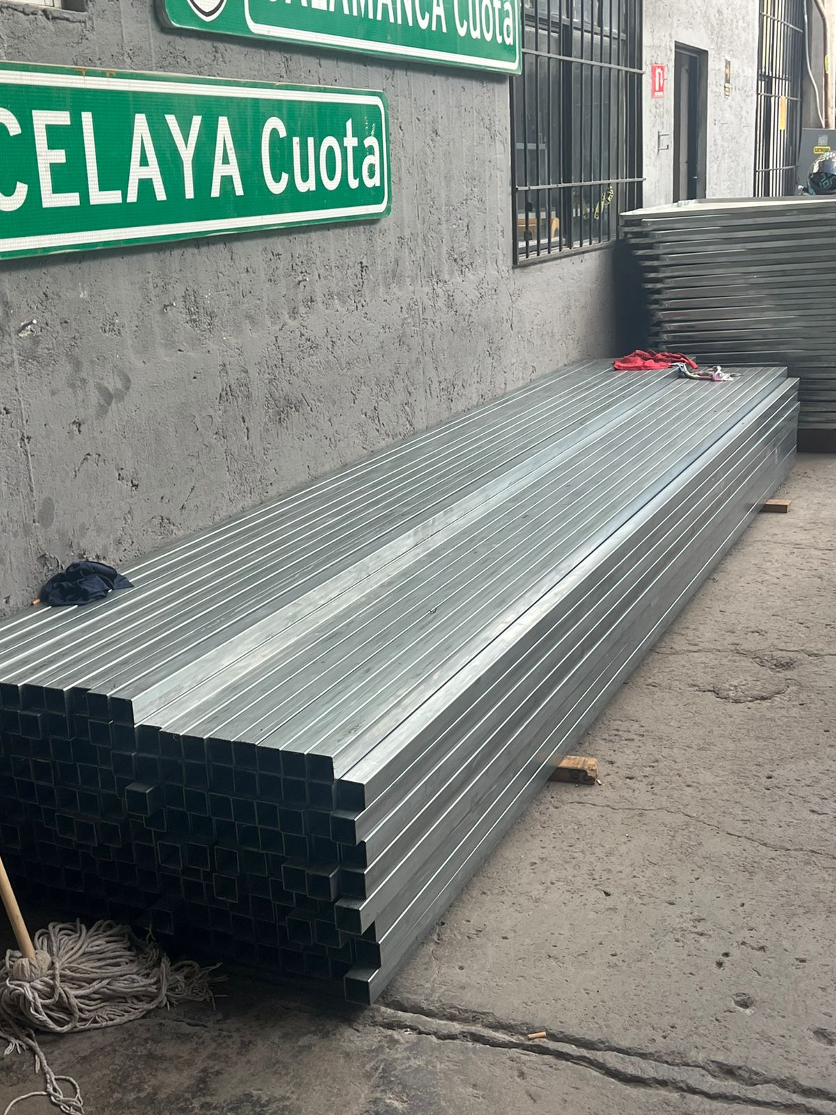 Tuberías de metal apiladas en el suelo junto a señales verde de Celaya Cuota y Salamanca Cuota en una pared gris.