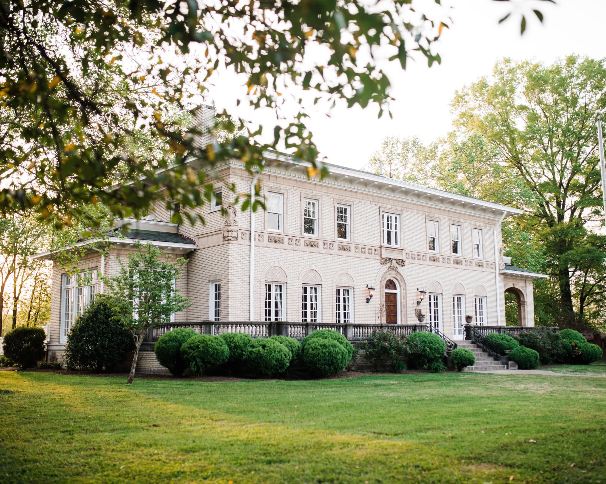 Elegant white brick mansion surrounded by lush green lawn and trees at sunset.