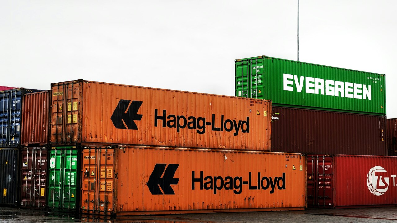 Contenedores apilados de empresas navieras Hapag-Lloyd y Evergreen en un puerto.