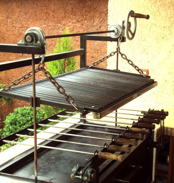 Parrilla de asado ajustable al aire libre con manivela y cadenas, sobre fondo de pared de ladrillo y vegetación.