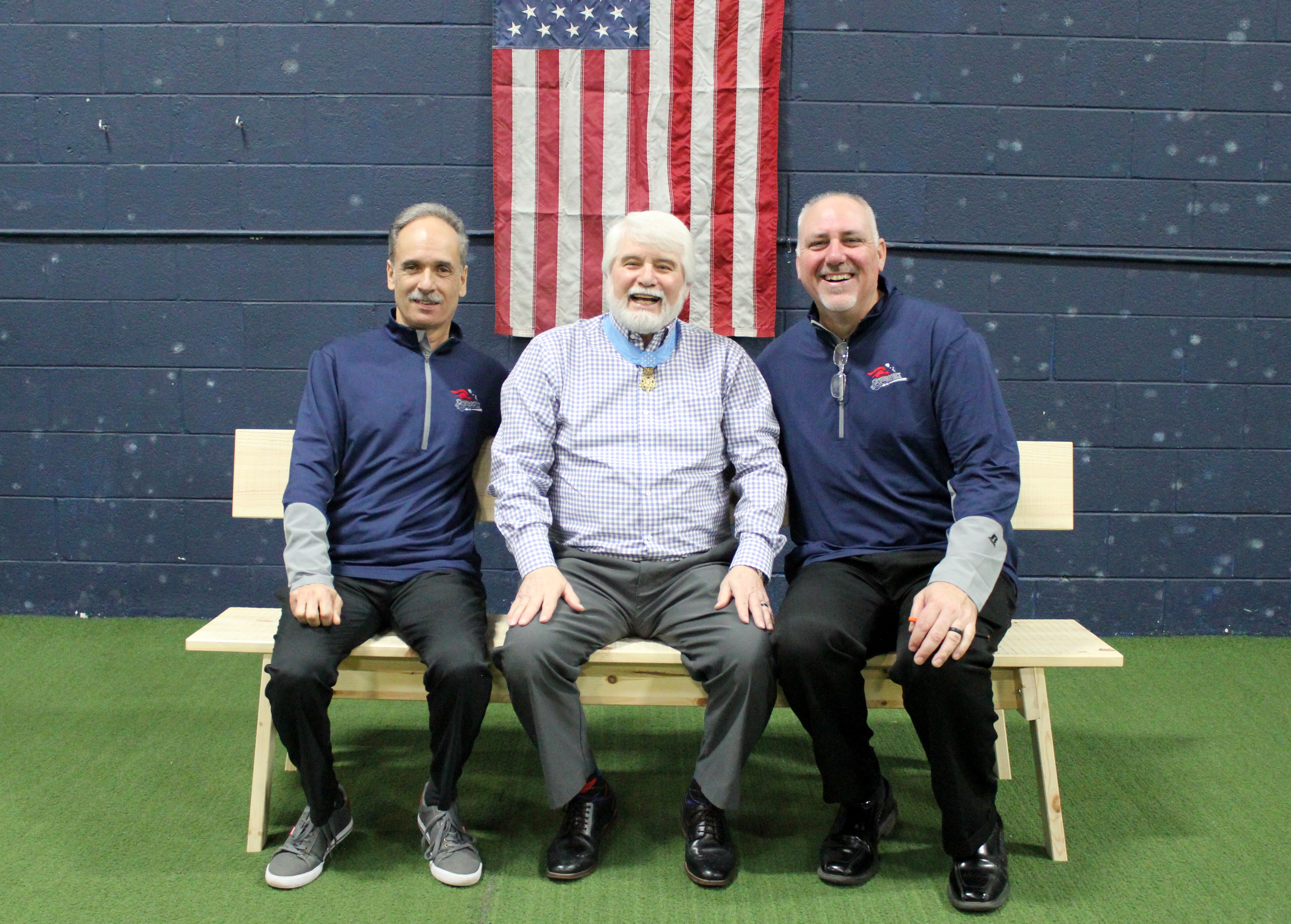Three men seated on a wooden bench in front of a wall with an American flag, wearing matching navy jackets and smiling.