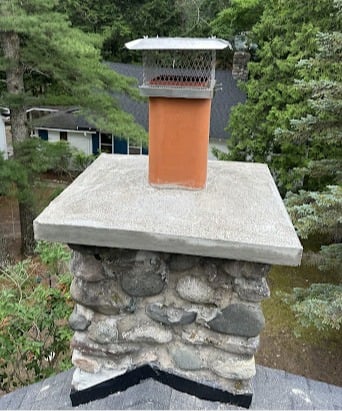 Stone chimney with metal cap on rooftop surrounded by lush green trees.
