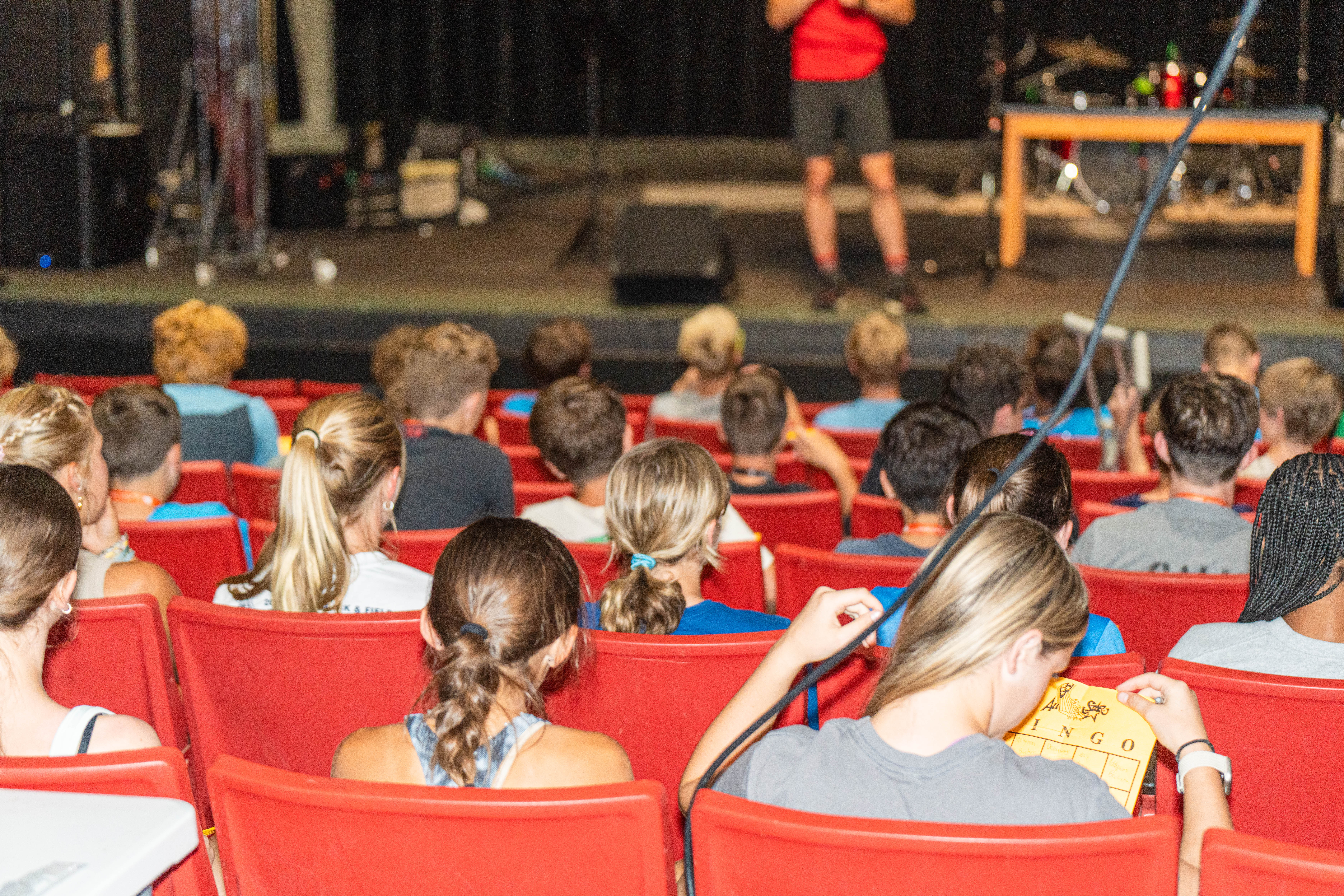 Audience seated in red chairs watching a presentation on stage, focus on participants engaged with bingo cards, in a large auditorium setting.