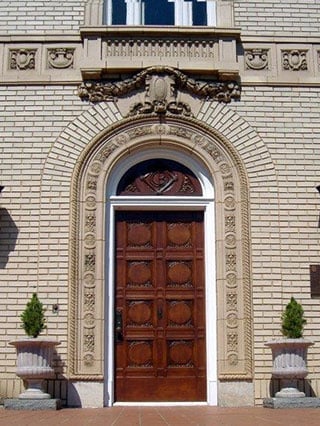 Intricately carved wooden door set in ornate brick archway with decorative stone elements and flanked by potted plants.
