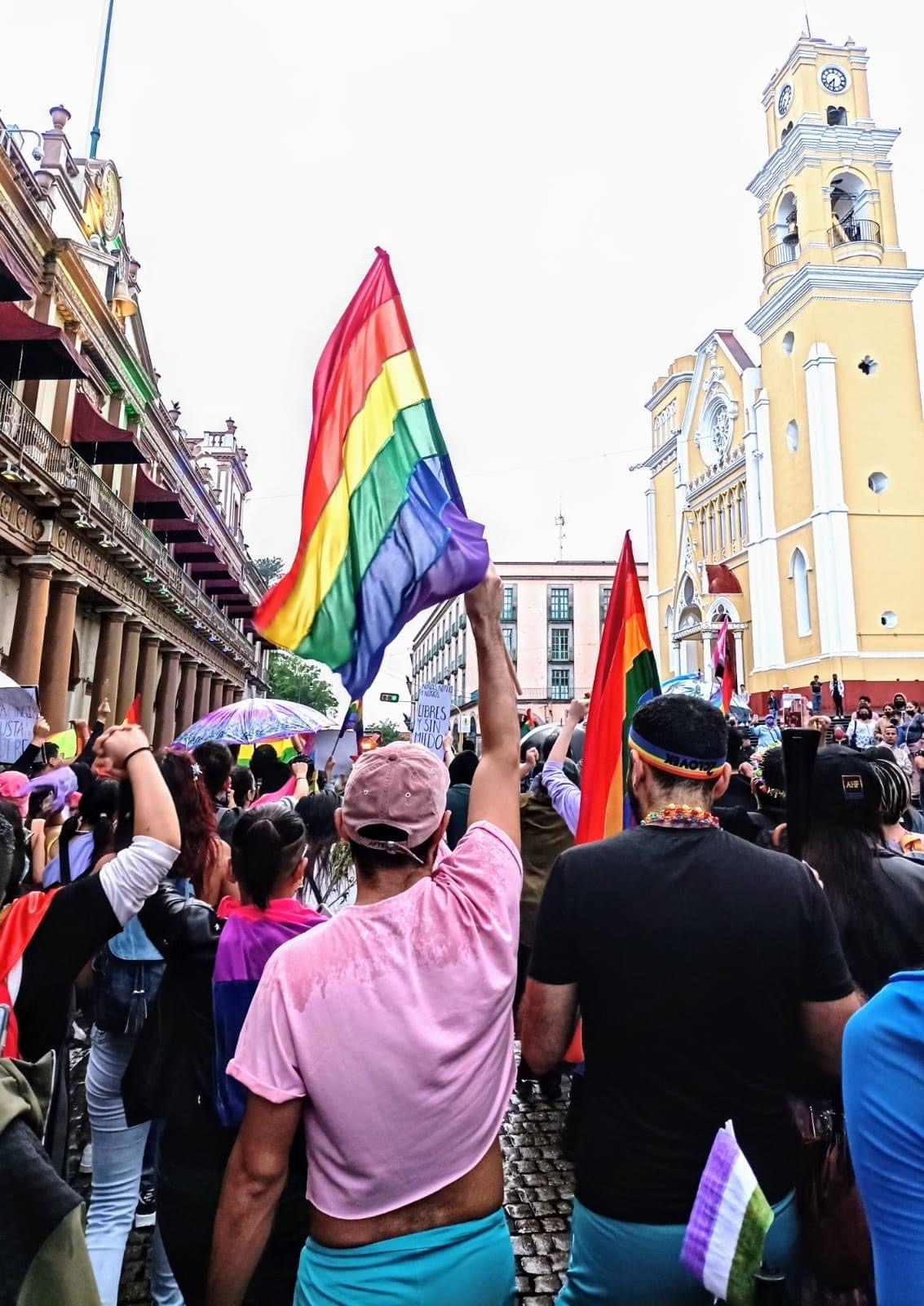 Manifestación del orgullo LGBTQ+ con una multitud marchando, banderas arcoíris ondeando y una iglesia amarilla de fondo.