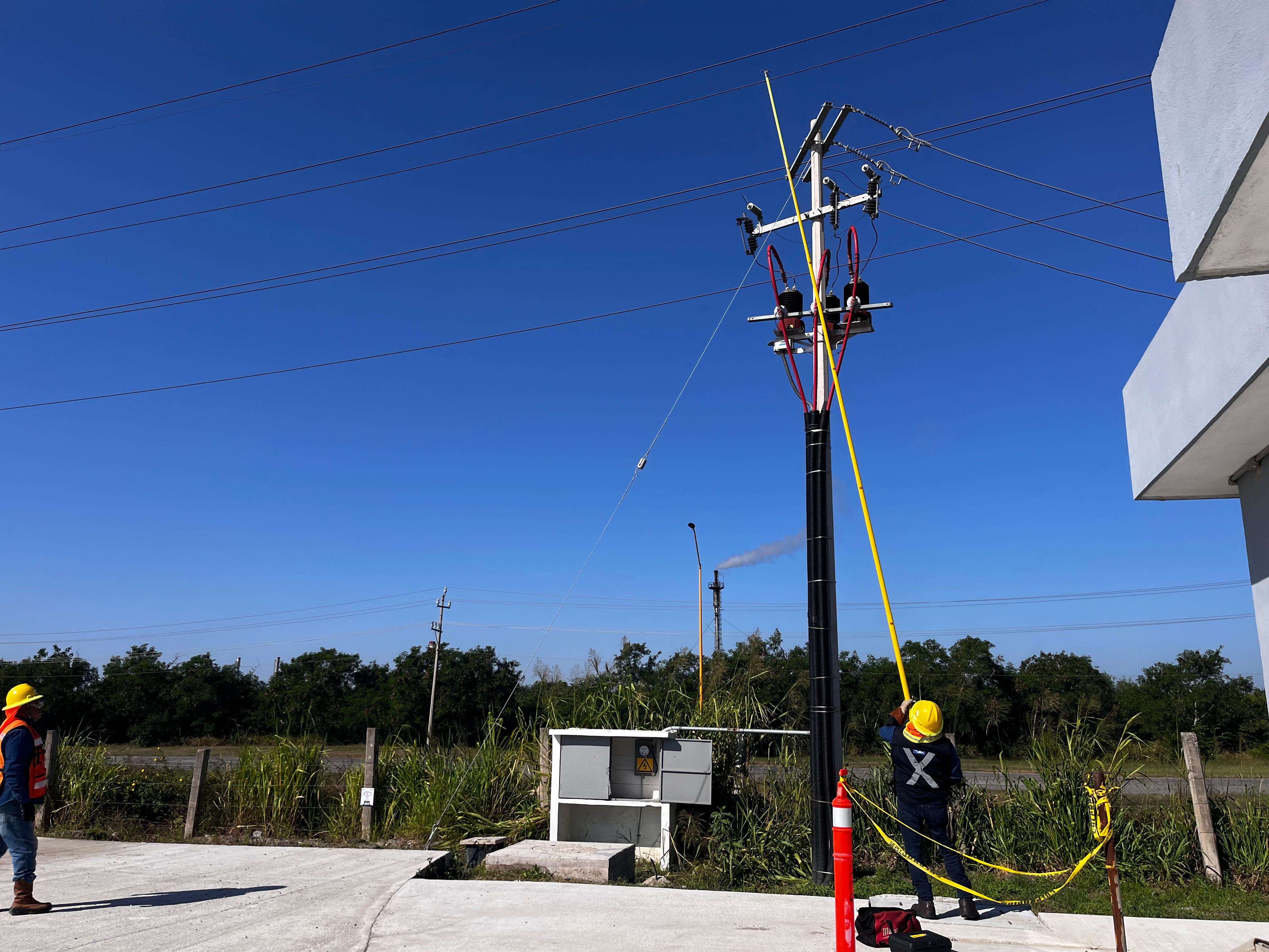 Trabajadores de mantenimiento con cascos amarillos utilizando herramientas largas para reparar líneas eléctricas en un día soleado.