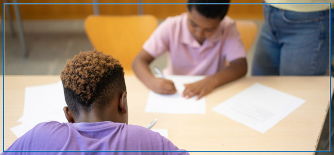Two students at a table doing school word with a Teacher in the background