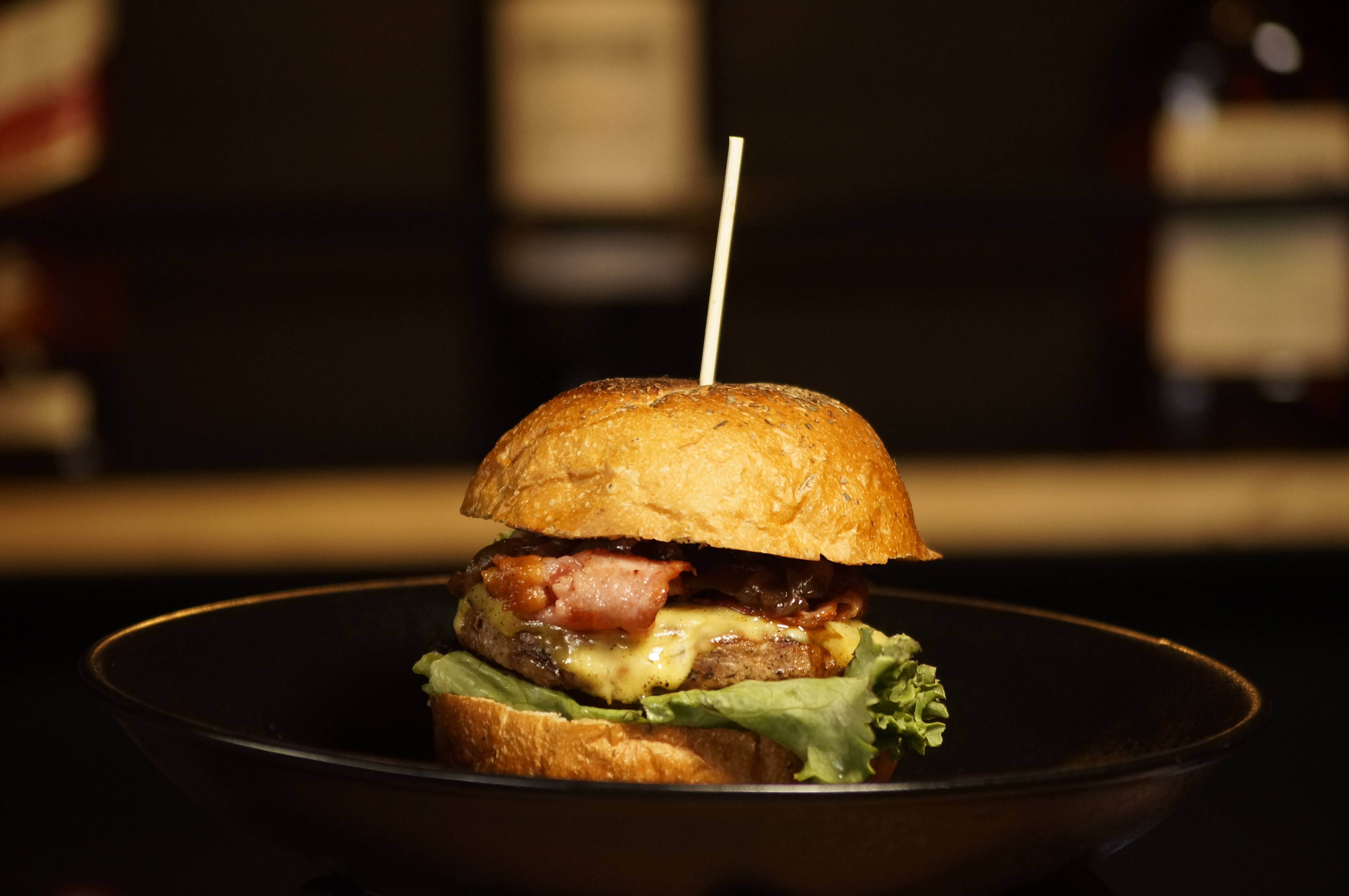 Close-up of a gourmet burger with lettuce, cheese, and bacon on a dark plate against a blurred background.