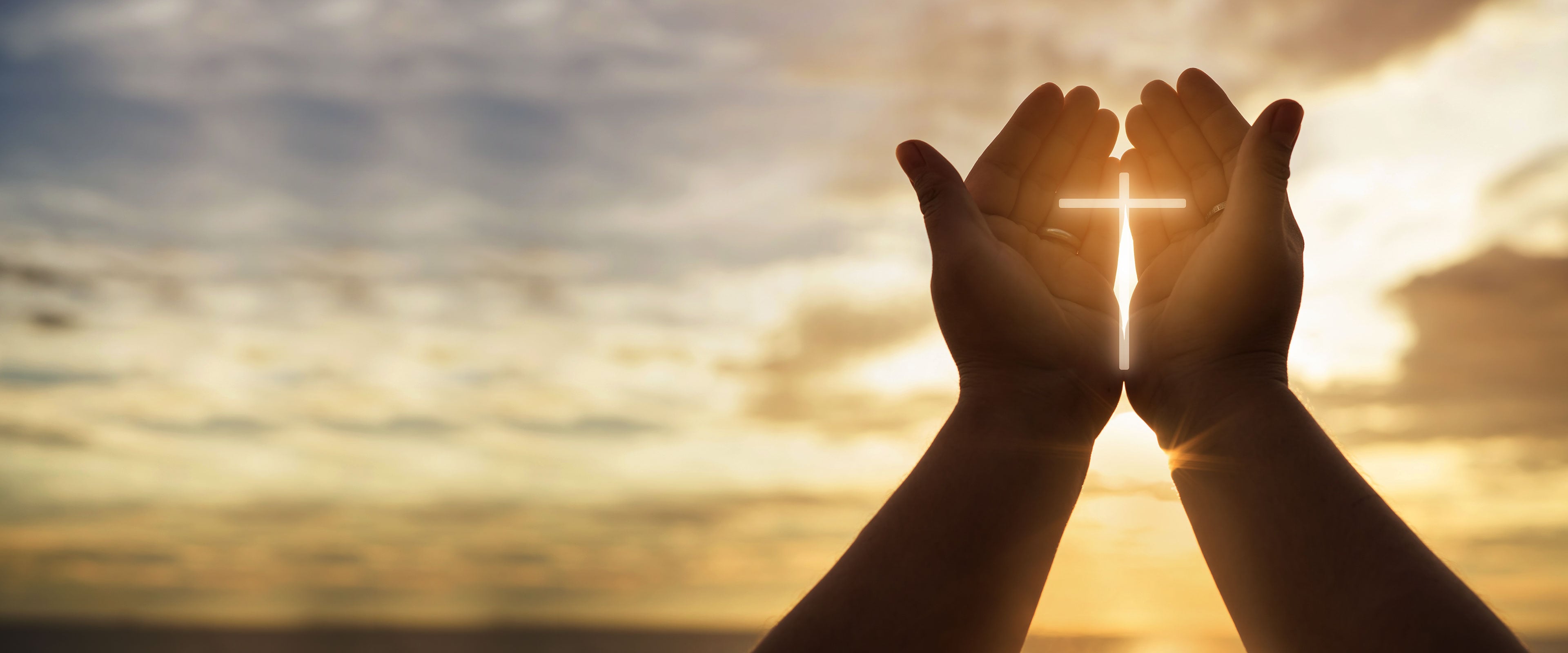 Hands holding a glowing cross against a sunset sky background, symbolizing faith and spirituality.