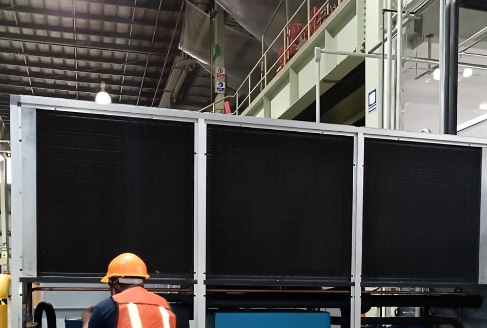 Trabajadores con cascos naranjas supervisando la instalación de un equipo industrial grande con grúa bajo cielo azul, con texto 'Ver Servicio Instalaciones'.