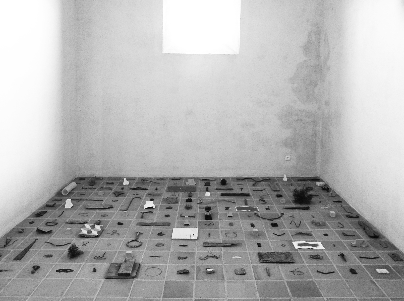 Objects arranged in a grid on a tile floor in an otherwise empty, monastic space.